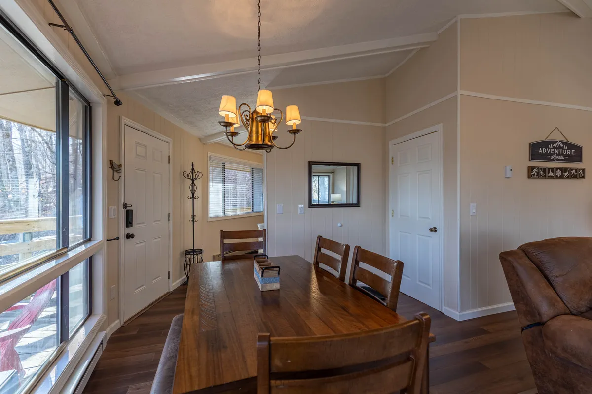 A dining area with a sturdy wooden table set beneath a stylish chandelier. Large windows surround the space, offering glimpses of the outdoors and inviting ample natural light. The layout suggests both casual family meals and festive gatherings.