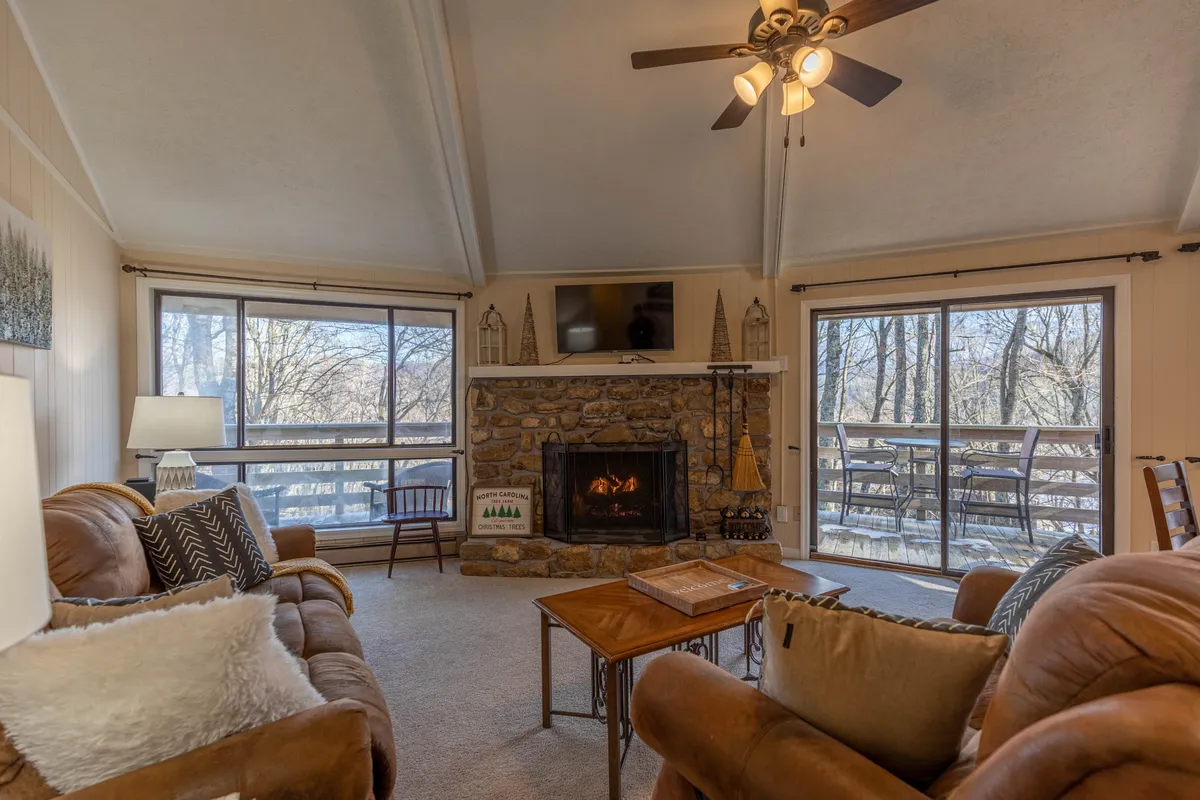 The hero image showcases a welcoming living area with a striking stone fireplace as its centerpiece, surrounded by plush, comfortable seating. Expansive windows fill the space with natural light and offer scenic views of Sugar Mountain, creating an atmosphere perfect for relaxation or social gatherings.