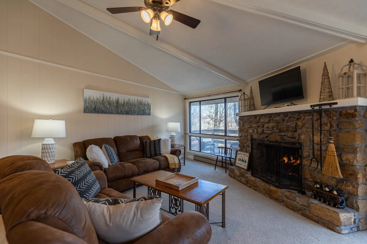 A comfortable brown sofa is arranged facing a stone fireplace, with a large window offering views of the outdoors. The inviting setup encourages relaxation and socializing, perfectly aligning with the room's purpose as a hub for unwinding after mountain adventures.