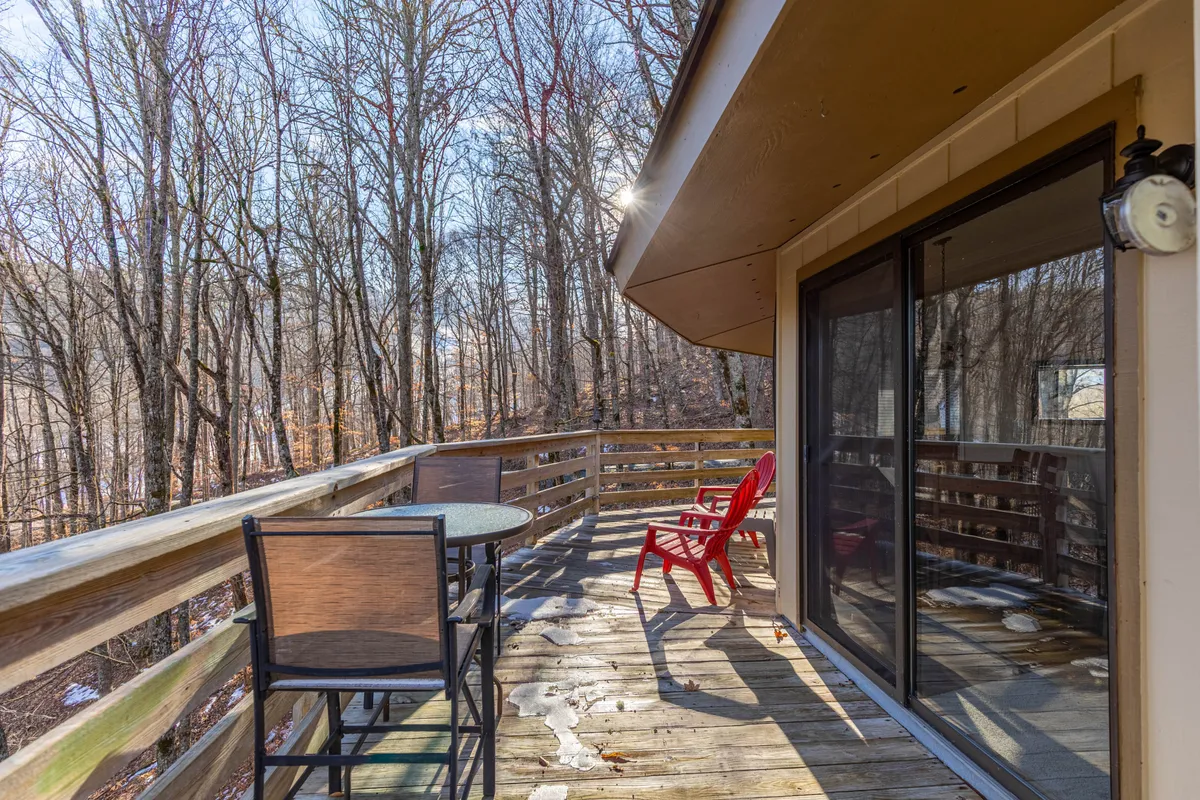 A spacious wooden deck with red chairs and a round table overlooks a lush treescape, evoking a serene spot for morning coffee, reading, or sharing moments with friends. The deck wraps around the exterior, emphasizing its use as a private perch for relaxation and gathering.