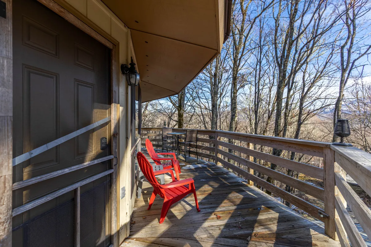 This image captures the cabin's entrance door framed by a wooden deck, with bright red chairs positioned for comfortable seating and a broad view of the natural landscape beyond. The wooden railing and the inviting approach to the doorway signal a welcoming transition from the outdoors to the interior space.