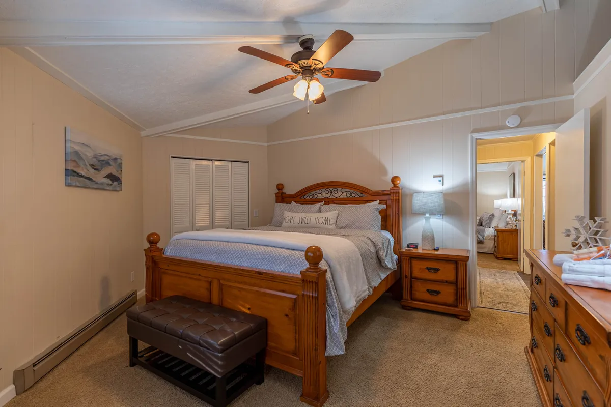 A sunlit bedroom with a neatly made bed, a nightstand, a comfortable bench, and calming wall art. Natural light fills the space, highlighting its serene and welcoming atmosphere—ideal for restful family downtime.