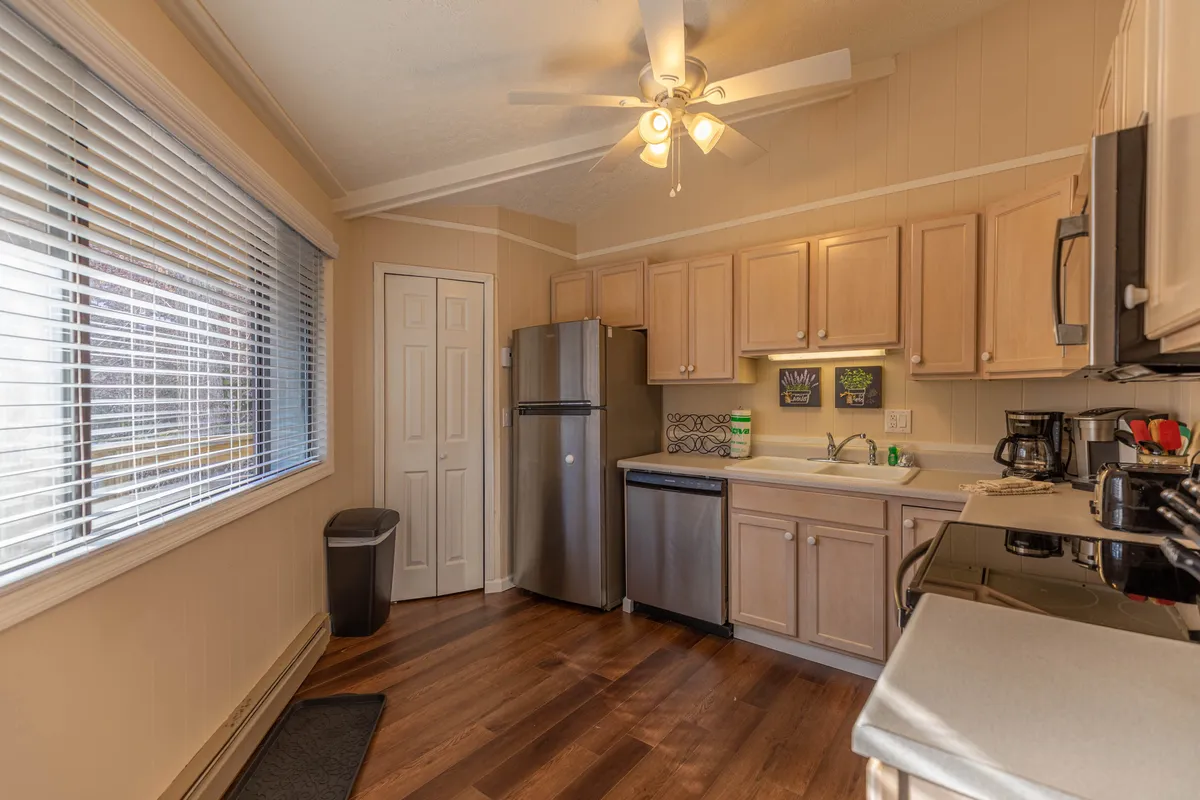 A warm, inviting kitchen featuring modern appliances, wooden cabinets, and a large window that fills the space with natural light. The countertops are clear and ready for meal preparation, with a ceiling fan overhead and enough space for several people to gather and cook together.