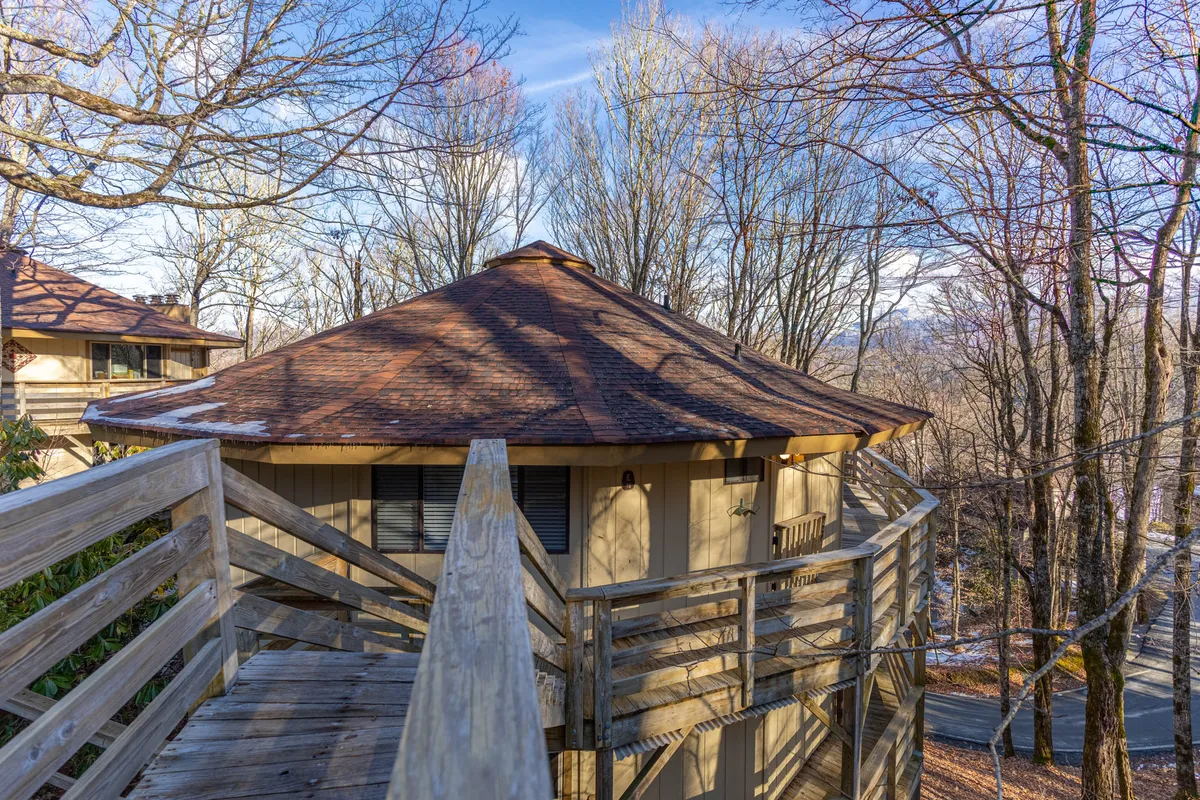 This image features the exterior of Sugar Mountain Serenity, focusing on a distinctive circular roof structure and a wooden walkway surrounded by lush trees. The scene embodies a tranquil mountain retreat, inviting guests to enjoy the natural surroundings from the comfort of the deck.