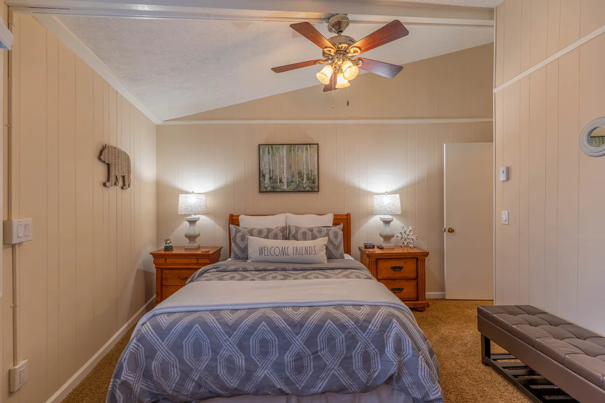 A cozy, inviting bedroom scene featuring a comfortable bed adorned with pillows and soft linens, flanked by bedside lamps and tranquil artwork. A chair in the corner offers a spot for reading or relaxing, while warm lighting creates a welcoming ambiance.