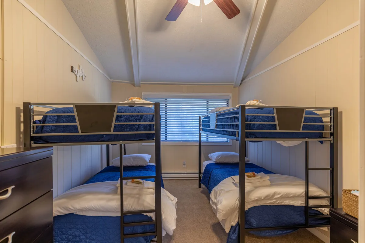 A playful bunk room with two sets of bunk beds, a window with blinds letting in natural light, and a ceiling fan for comfort. The vibe is youthful and relaxed, perfect for families or kids to enjoy games and late-night chats.