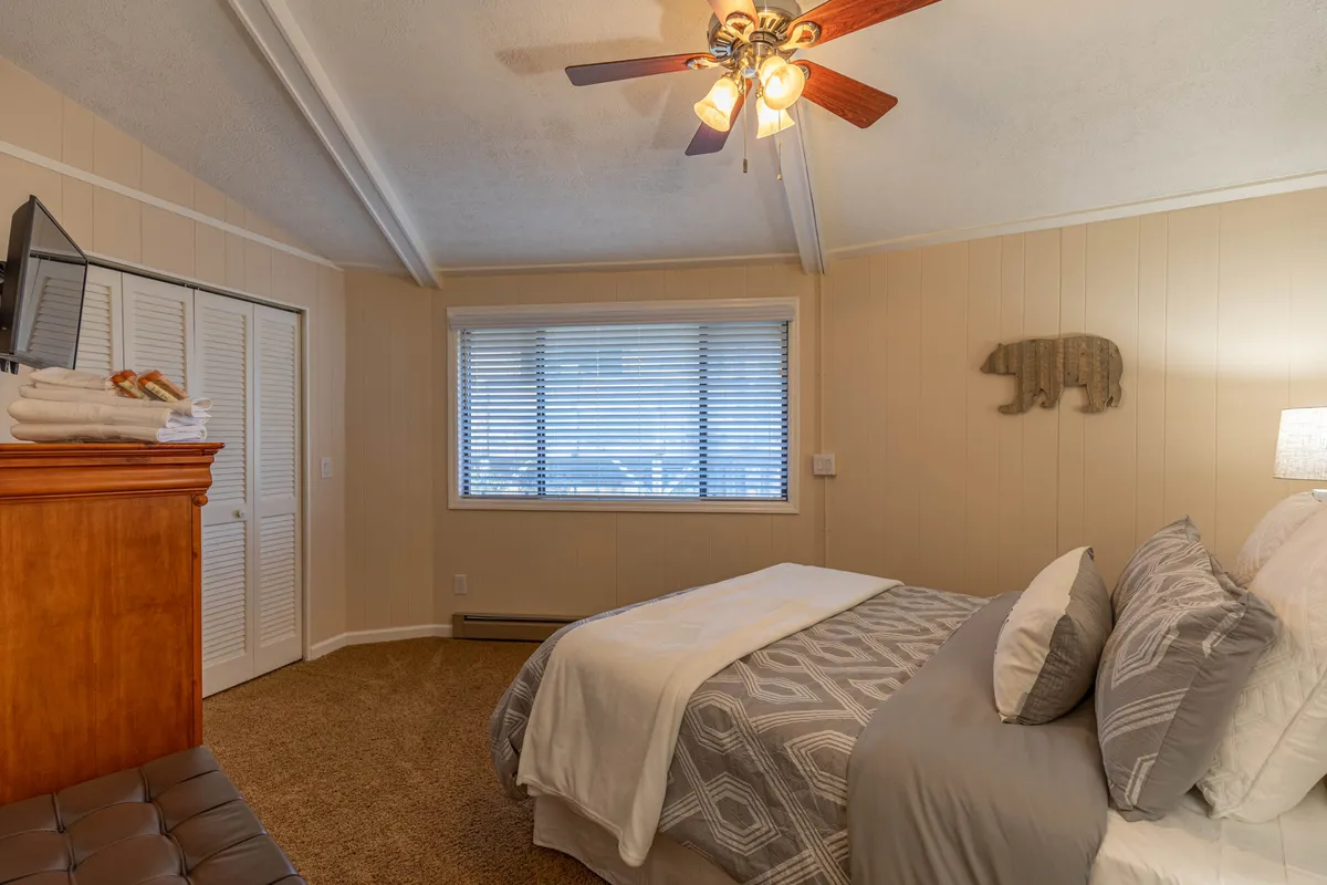 A spacious master bedroom with a plush king bed adorned in gray and white bedding. The wooden headboard adds warmth, while a bear-themed piece of wall art lends a playful, mountain-touch. Large windows allow natural light to flood the room, and a ceiling fan ensures comfort. There's a TV positioned for easy viewing from bed, creating an inviting atmosphere for relaxation and entertainment.
