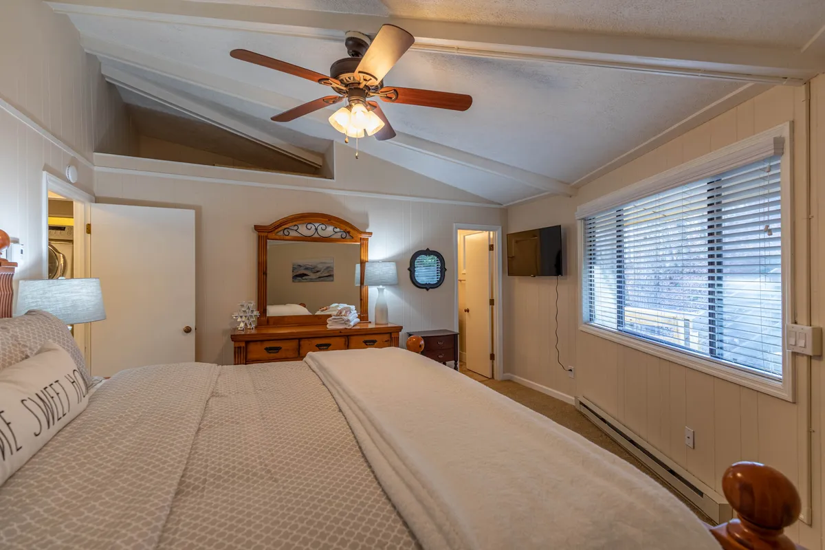 A comfortable queen bedroom featuring a bed with decorative pillows, a wooden dresser, and a large window with blinds. A ceiling fan hangs overhead, and the room is filled with soft, natural light, creating an atmosphere of relaxation and privacy.